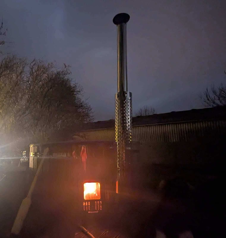 Nächtliche Außenszene: Leuchtender Holzofenkamin mit Hitzeschutzblech aus Metall neben kahlen Bäumen und dunkler Gebäudesilhouette.