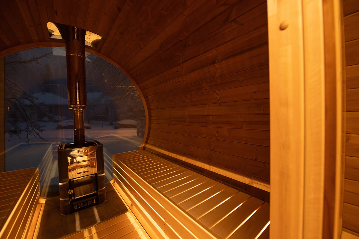 Barrel sauna with benches and a stove, surrounded by snow outside.