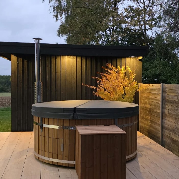 Abendliche Außenansicht eines geschlossenen, runden Holz-Whirlpools mit Überdachung in der Nähe eines schwarzen Schuppens und eines beleuchteten Baums.