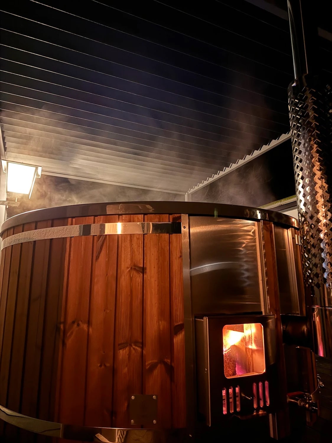 Nachtansicht eines holzbefeuerten Hot Tubx mit durch das Ofenfenster sichtbarem Feuer und aufsteigendem Dampf unter dem überdachten Außenbereich