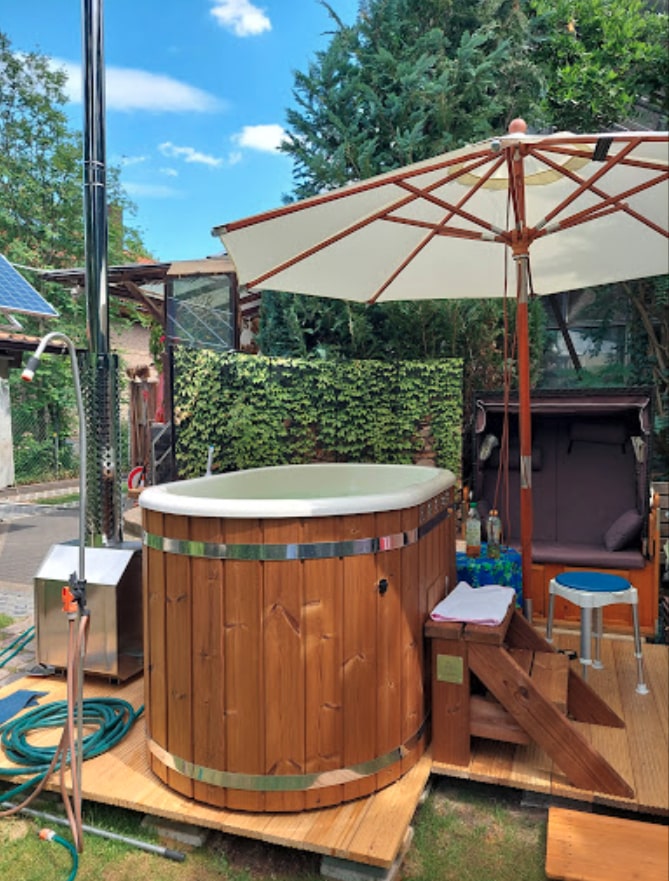 Außenansicht eines hölzernen Whirlpools mit Metallbändern, umgeben von einem Sonnenschirm, Holzstufen und Gartengrün im Hintergrund