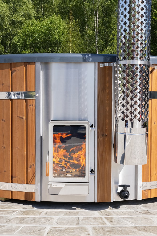 Integrated stainless steel wood-fired hot tub heater with glass fire viewing window and chimney connection.