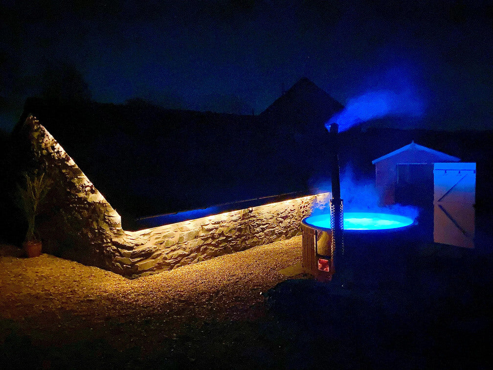 Nächtliche Außenszene mit einem leuchtend blauen Whirlpool neben einer Steinmauer und einem Schuppen in Dunkelheit