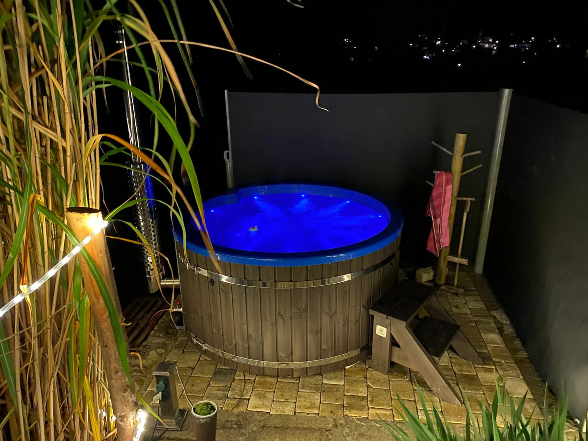 Nachtansicht eines runden hölzernen Whirlpools mit blauer LED-Beleuchtung auf einer Steinterrasse, kleinen Holzstufen und Außendusche