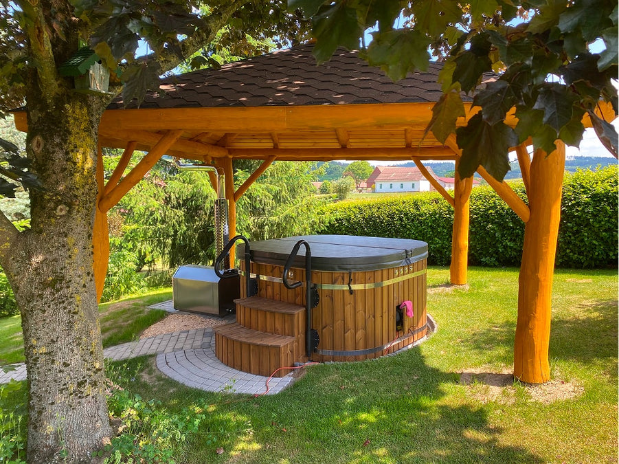 Außenansicht eines hölzernen Whirlpools mit Treppe und Überdachung unter einem Pavillon, umgeben von üppigem Grün und Bäumen
