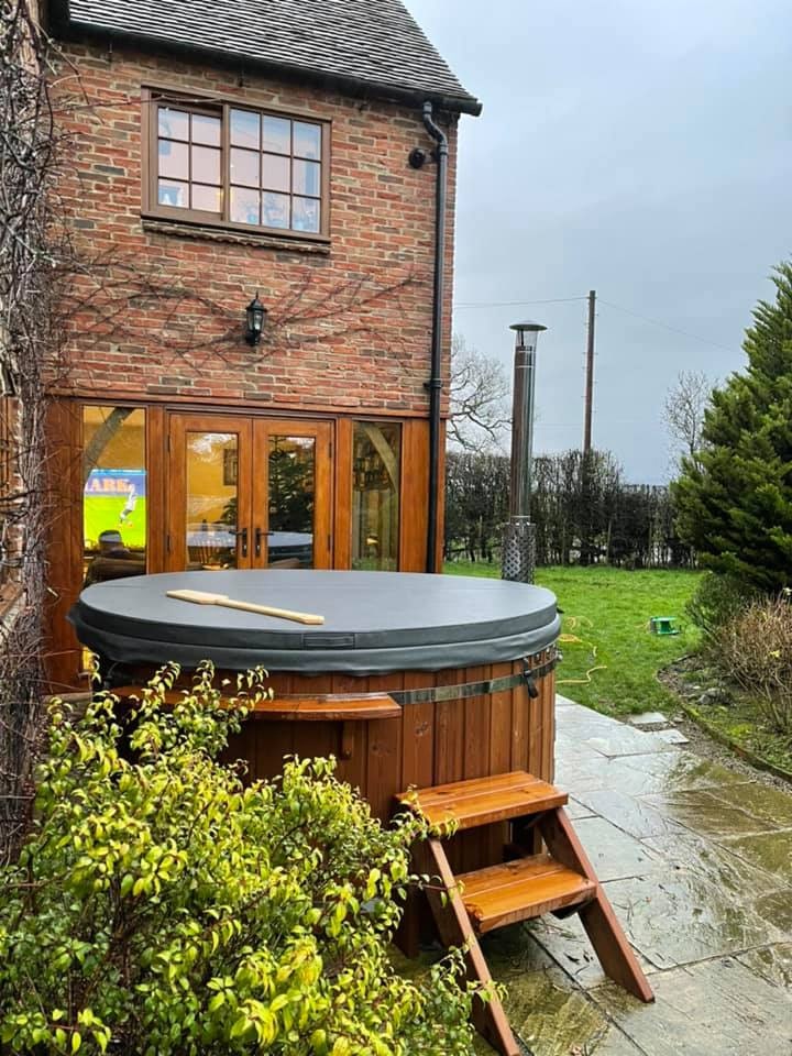 Ein holzbefeuerter Hot Tubx von Gardenvity wurde vor einem Backsteinhaus mit Rasen und Blick auf die Landschaft installiert.