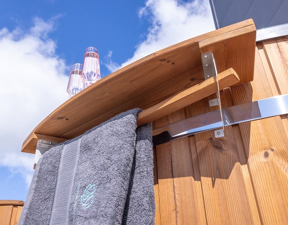 Nahaufnahme eines hölzernen Handtuchhalters, der an einem holzbefeuerten Whirlpool befestigt ist und zwei graue Handtücher trägt, vor blauem Himmel.