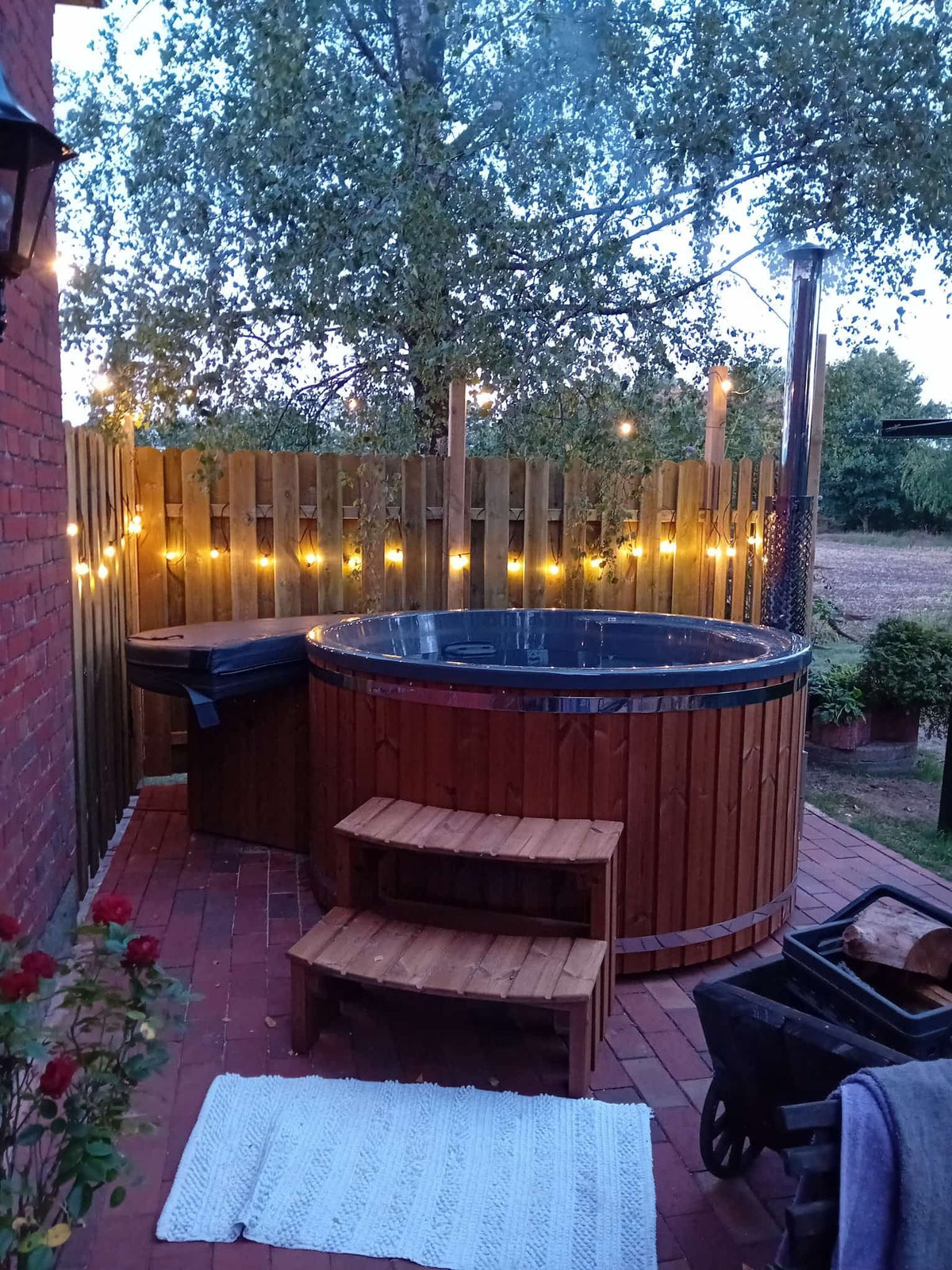 Außenansicht eines hölzernen Whirlpools mit zweistufiger Holztreppe, Lichterketten am Zaun und Backsteinterrasse in der Abenddämmerung