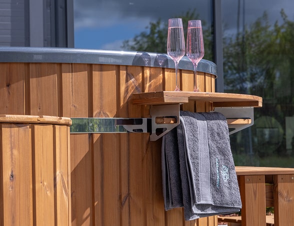 Nahaufnahme der hölzernen Seite des hot tub mit klappbarem Tablett, auf dem zwei Gläser und ein hängendes dunkelgraues Handtuch mit gesticktem Logo zu sehen sind.