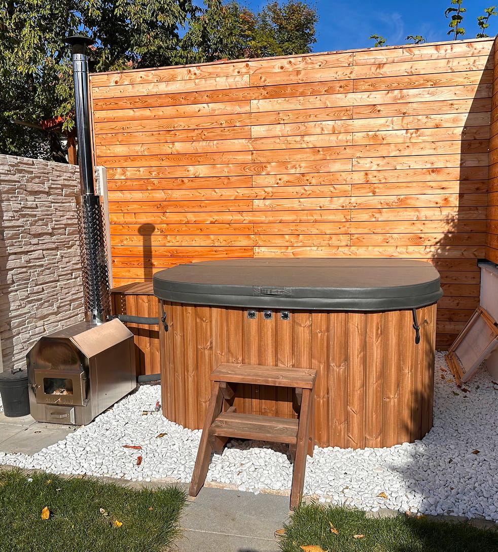Quadratischer Whirlpool mit Abdeckung und Standardstufe in einer Gartenanlage mit Holzzaun und Bäumen.